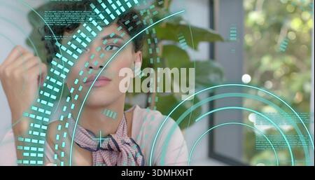 Gesturing adult man wearing light top and patterned scarf using glass panel, teal HUD, potted plant Stock Photo