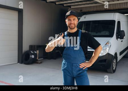 Thumb up, positive facial expression. Delivery man is by the bus near garage outdoors. Stock Photo