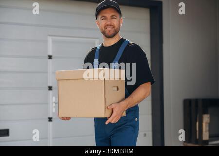 Mailing delivery service worker is in blue uniform, holding box. Stock Photo