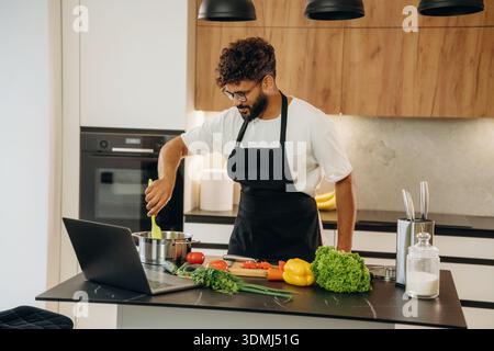 Taking online lessons, using laptop. Handsome man is preparing food on the kitchen. Stock Photo
