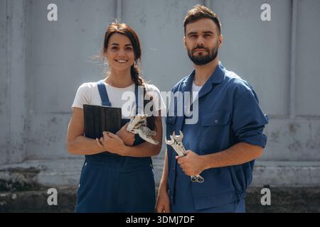 Holding tools and digital tablet. Cheerful male and female mechanics are standing outdoors together. Stock Photo