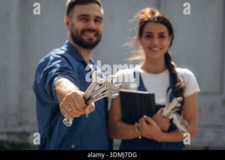 Holding tools and digital tablet. Cheerful male and female mechanics are standing outdoors together. Stock Photo