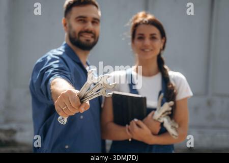 Holding tools and digital tablet. Cheerful male and female mechanics are standing outdoors together. Stock Photo
