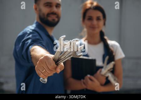 Holding tools and digital tablet. Cheerful male and female mechanics are standing outdoors together. Stock Photo