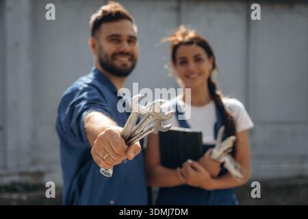 Holding tools and digital tablet. Cheerful male and female mechanics are standing outdoors together. Stock Photo