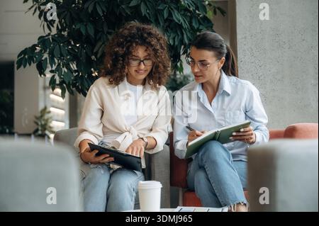 Sitting on the sofa with notepad. Two women are indoors in the modern hall of building at daytime. Stock Photo
