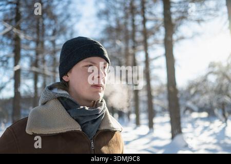 Close portrait of young man in winter clothing exhaling visible breath in cold snowy park. Concept of cold weather and winter lifestyle. Stock Photo