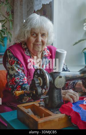 An elderly woman looks at the camera while using a sewing machine Stock Photo