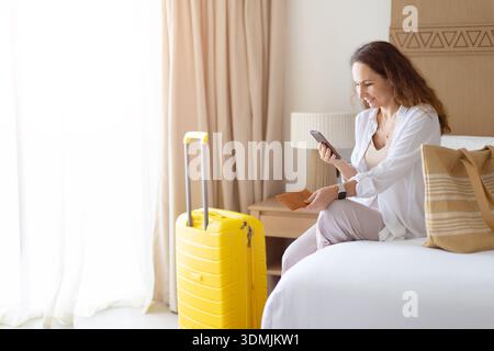 Adult woman sitting on bed, using smartphone near suitcase, preparing for travel Stock Photo