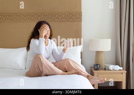 Woman at home sitting on bed holding glass of water, feeling unwell Stock Photo