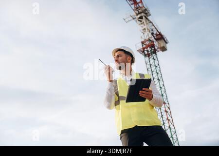 Controlling the process, with walkie talkie. Construction site worker is against building crane and sky background. Stock Photo