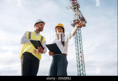 Man and woman, construction site workers are against crane, communication. Stock Photo