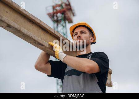 A construction worker is moving the wooden plank. In the yellow hard hat and in gloves. Stock Photo