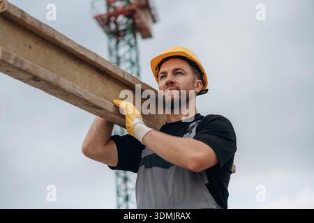 A construction worker is moving the wooden plank. In the yellow hard hat and in gloves. Stock Photo