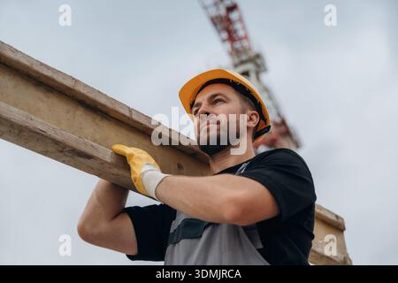 A construction worker is moving the wooden plank. In the yellow hard hat and in gloves. Stock Photo