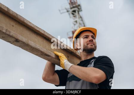 A construction worker is moving the wooden plank. In the yellow hard hat and in gloves. Stock Photo