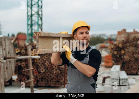 Looking to the side, moving the wooden plank. A builder is working at the construction site at daytime. Stock Photo