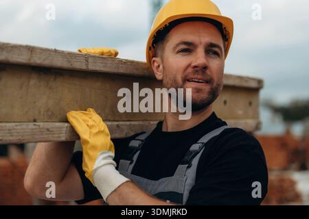 Looking to the side, moving the wooden plank. A builder is working at the construction site at daytime. Stock Photo