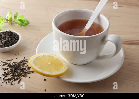 Background with detail of black tea with lemon in a white ceramic cup on a wooden table and dried black tea leaves. Elevated view. Stock Photo