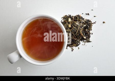 Detail of a white ceramic cup filled with hot tea on a white table with dried tea leaves. Top view. Stock Photo
