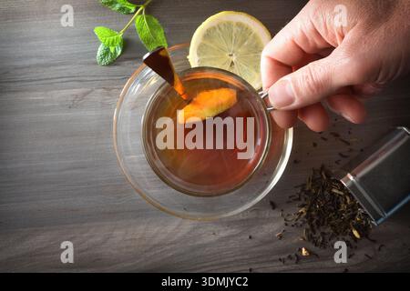 Person holding a cup of hot tea on a dark table with a metal container containing dried tea leaves, a slice of lemon, and a fresh mint leaf. Top view. Stock Photo