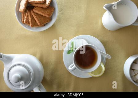 Tea infusion in a white ceramic cup with teapot, milk jug, sugar cubes and biscuits, in elegant white ceramic containers on a table with a beige fabri Stock Photo
