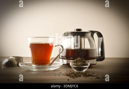 Glass cup and teapot full of tea on a wooden bench with a bowl of dried tea leaves with white isolated background. Front view. Stock Photo