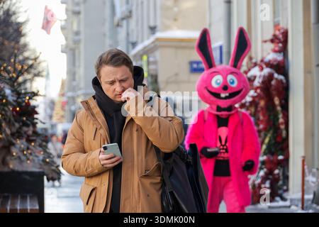 Man using smartphone on winter city street standing on background of person in pink bunny costume Stock Photo