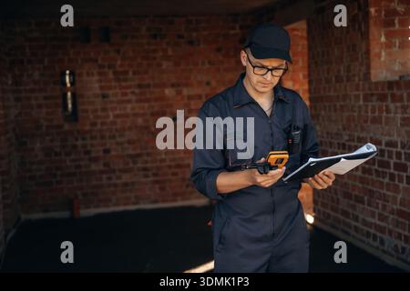 Documents and leveler tool are in hands. Builder is working on construction site, unfinished building. Stock Photo