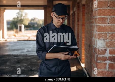 Documents and leveler tool are in hands. Builder is working on construction site, unfinished building. Stock Photo
