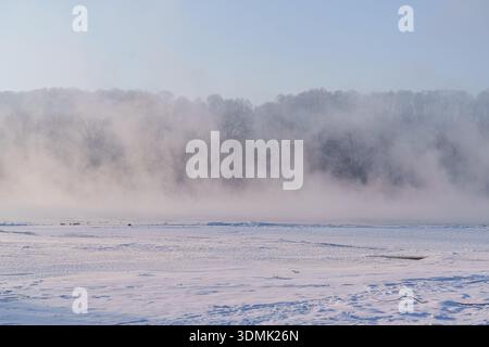 Thick fog and mist rising from a frozen river in a winter forest landscape. Stock Photo