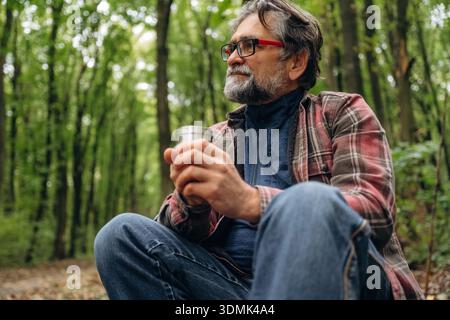Enjoying the hot drink from thermos. Hiking experience, senior man is in the forest at daytime. Stock Photo