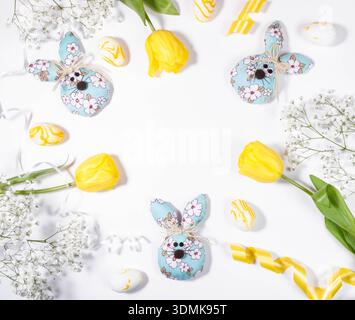Frame of three decorative Easter bunnies made of fabric with floral patterns are arranged with yellow tulips and colorful eggs on white background, cr Stock Photo