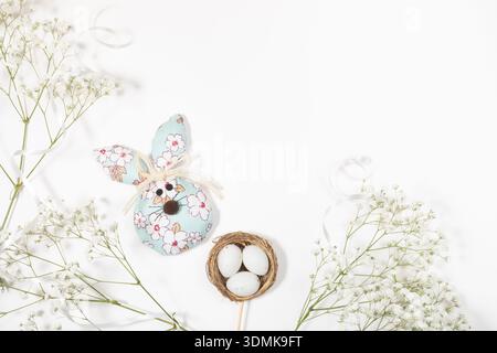 Decorative Easter bunny figure with floral patterns sits beside small nest containing white eggs, surrounded by delicate white flowers on white backdr Stock Photo