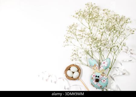 Decorative Easter bunny made of painted material sits beside nest containing three eggs, surrounded by delicate white flowers on clean white backgroun Stock Photo
