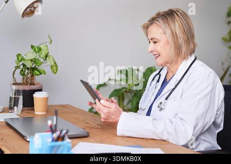 Friendly Female Doctor Using Smartphone At Desk For Telemedicine Consultation In Clinic Stock Photo
