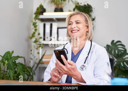 Smiling Female Doctor Using Smartphone In Clinic Office With Stethoscope Stock Photo