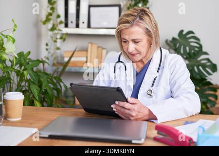 Dedicated Doctor Analyzing Tablet At Desk In Clinic Office With Plants Stock Photo