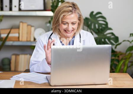 Smiling Female Doctor Waving Hello While Working on Laptop in Office Stock Photo