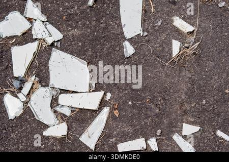 A top-down shot of several sharp, clear glass fragments and debris lying on a rough, dark gray asphalt surface, suggesting an accident or vandalism Stock Photo