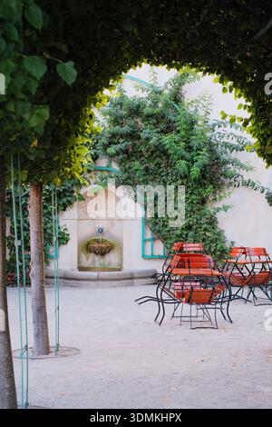 A tranquil garden scene featuring a historic stone wall fountain adorned with lush climbing ivy. The courtyard is arranged with elegant wooden cafe ch Stock Photo