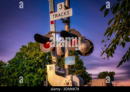 Active railroad crossing signal with red warning lights illuminated beneath a purple sunset sky near leafy trees and urban surroundings. Stock Photo