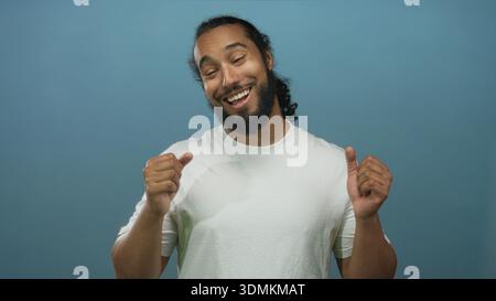 Man with hands over chest smiling in blue studio, wearing white t shirt and looking grateful; gratitude. Stock Photo