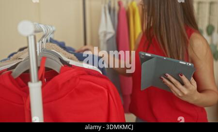 Woman holds tablet and touches clothes with manicured hands in a walkin closet studio wearing red top; thoughtful outfit choice. Stock Photo