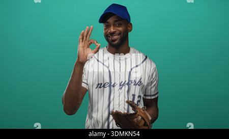 Man baseball player in pinstripe jersey and blue cap giving ok hand gesture with leather glove in studio; confidence. Stock Photo