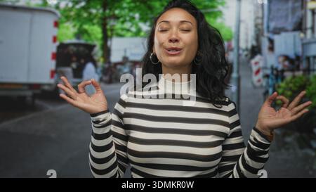 Woman blowing a kiss with hands visible on a city street wearing a black and white striped turtleneck and wooden watch; playful affection. Stock Photo