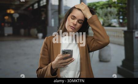 Hispanic woman holding smartphone looks distressed while standing outdoors in busy city street, showing signs of a headache under the daylight. Stock Photo