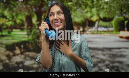 Woman holds blue telephone receiver to ear while hand rests over chest in park; gratitude connection. Stock Photo