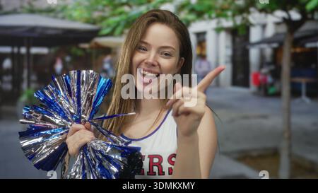 Young cheerleader woman holding blue metallic pompom points finger and smiles widely on street; enthusiasm. Stock Photo