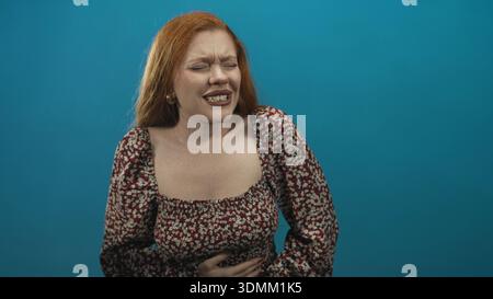 Woman clutching stomach, hands pressing abdomen, eyes closed and wincing in floral dress against teal backdrop in studio; pain. Stock Photo
